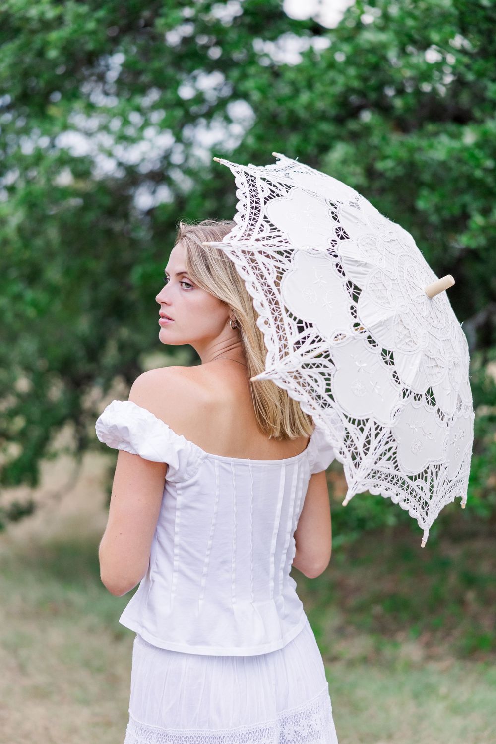 A person in a white dress holds a delicate lace parasol while standing in a lush green garden setting.