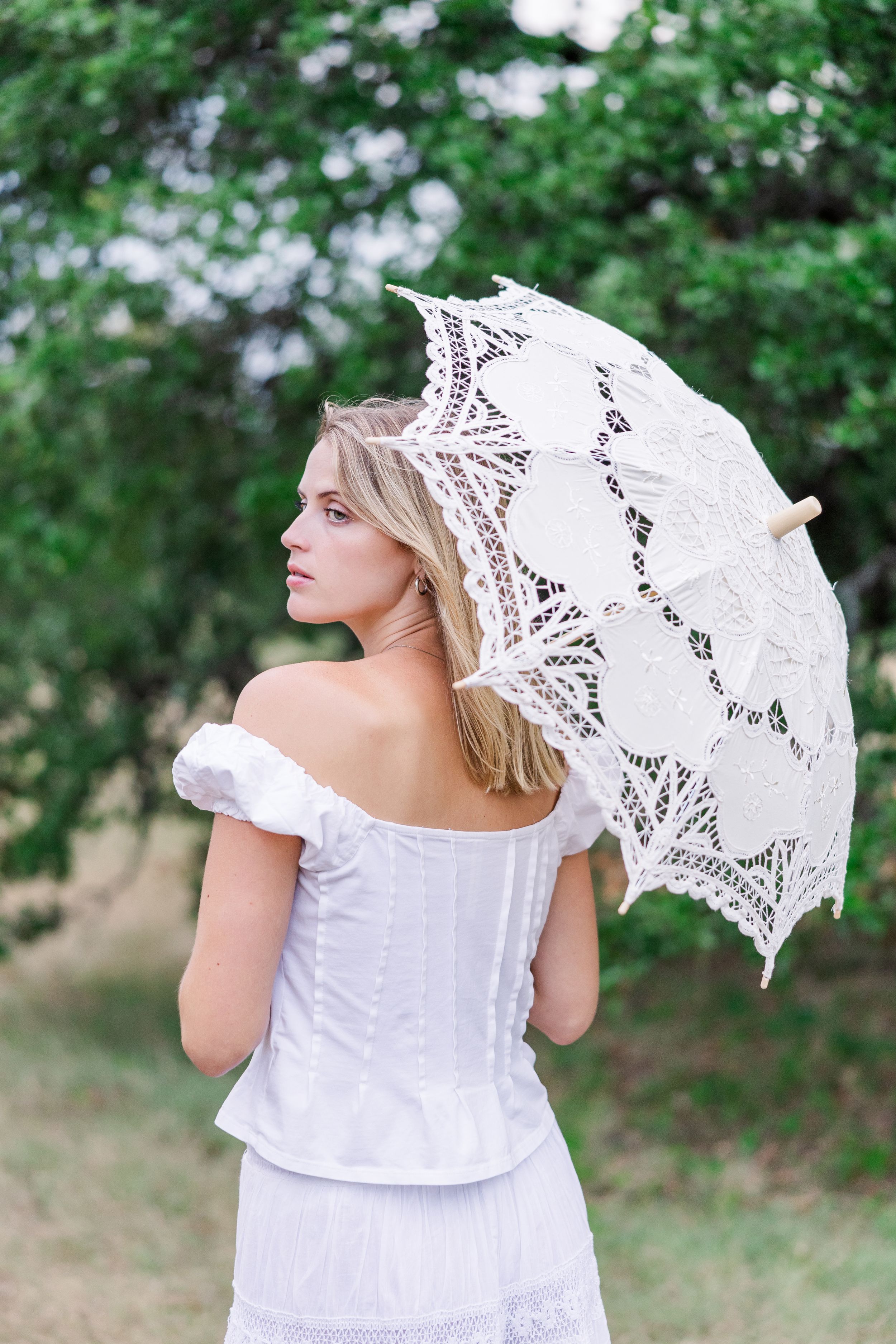 A person in a white dress holds a delicate lace parasol while standing in a lush green garden setting.