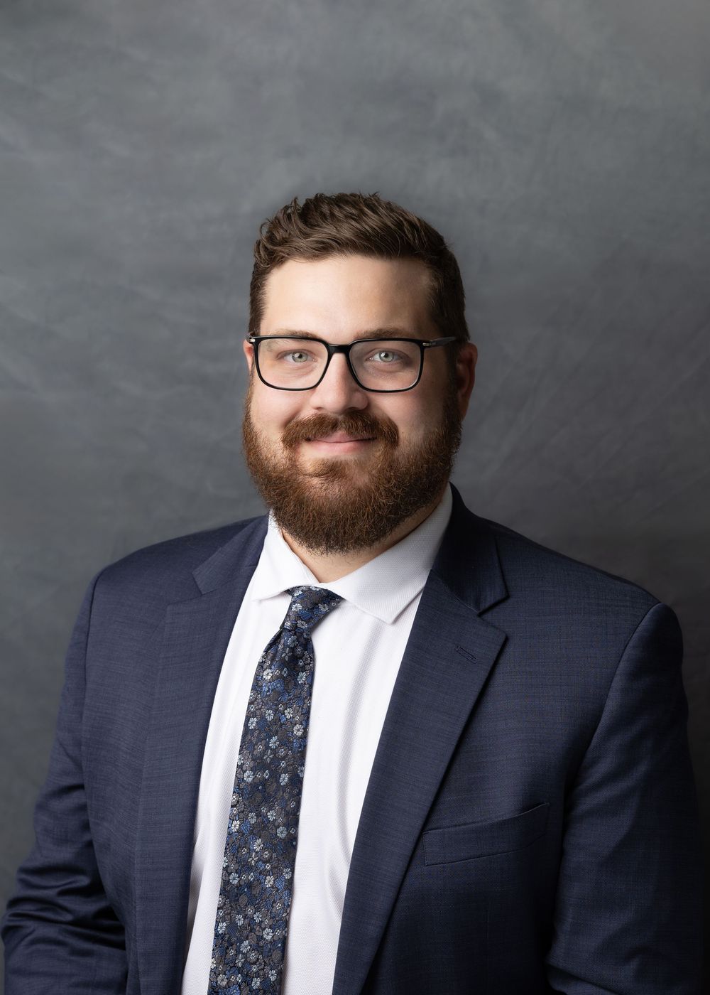 Professional headshot portrait against gray background showing person in navy suit with floral tie and glasses.