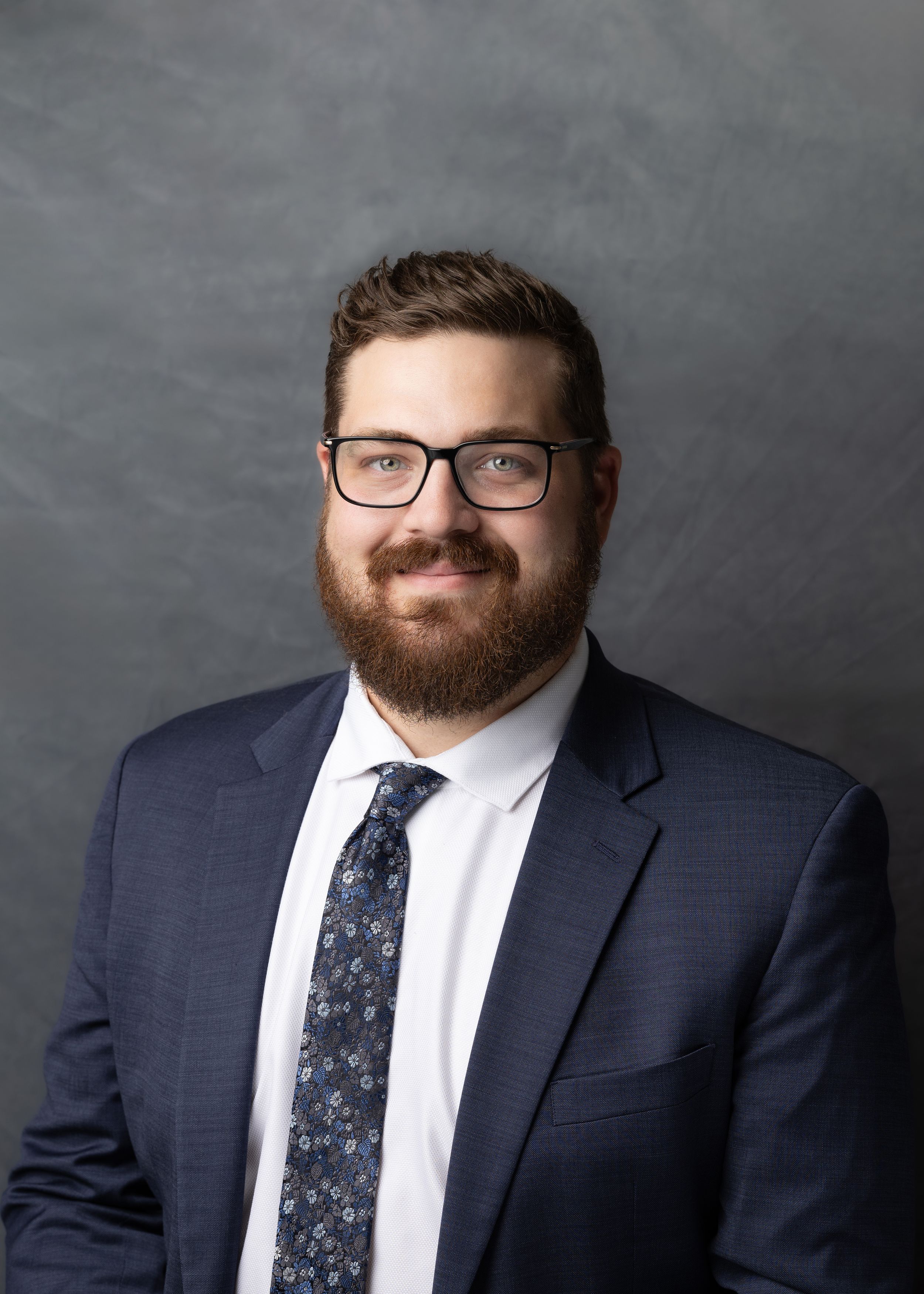 Professional headshot portrait against gray background showing person in navy suit with floral tie and glasses.