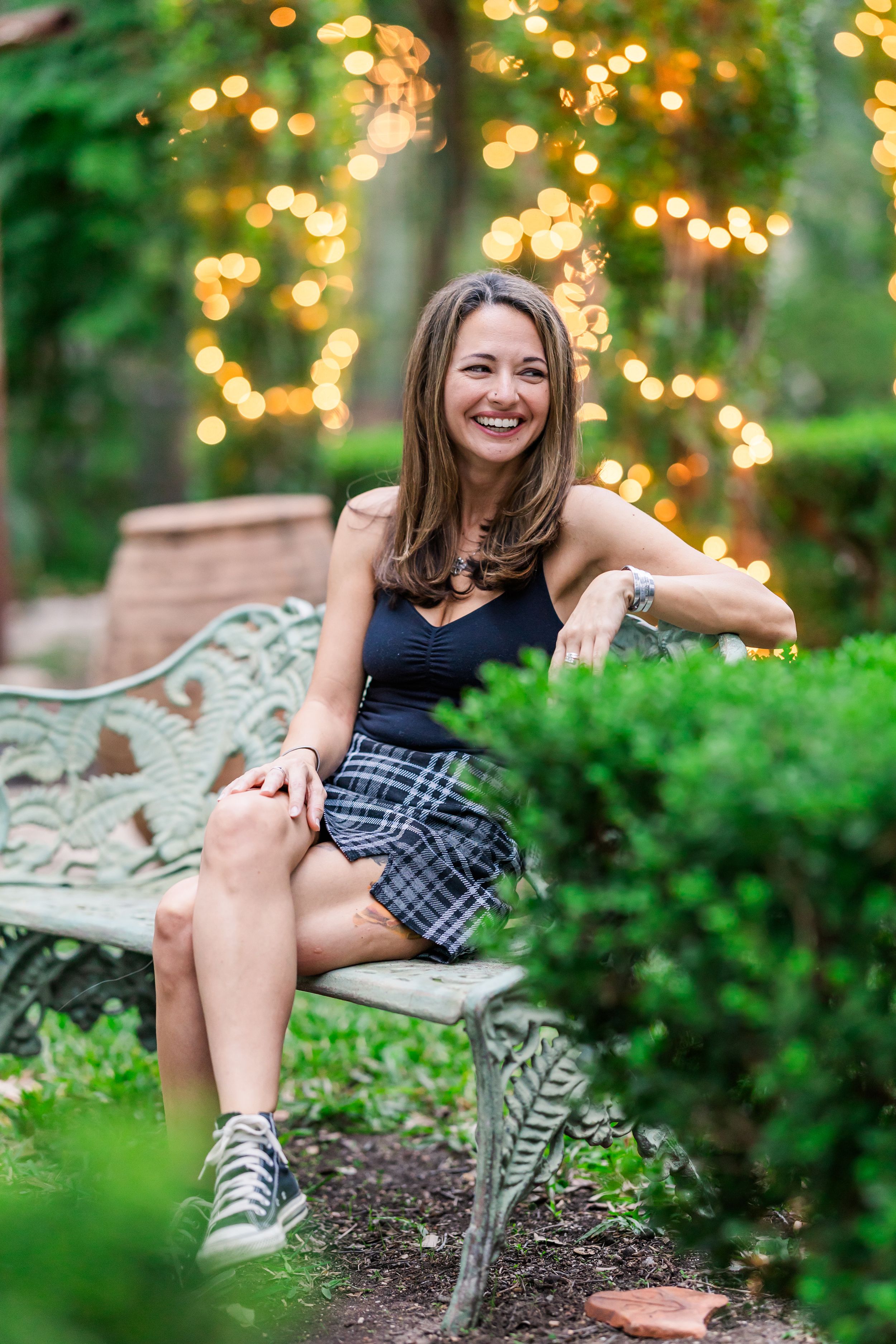 A person in a plaid dress and sneakers sits on a decorative garden bench with twinkling lights in the background.