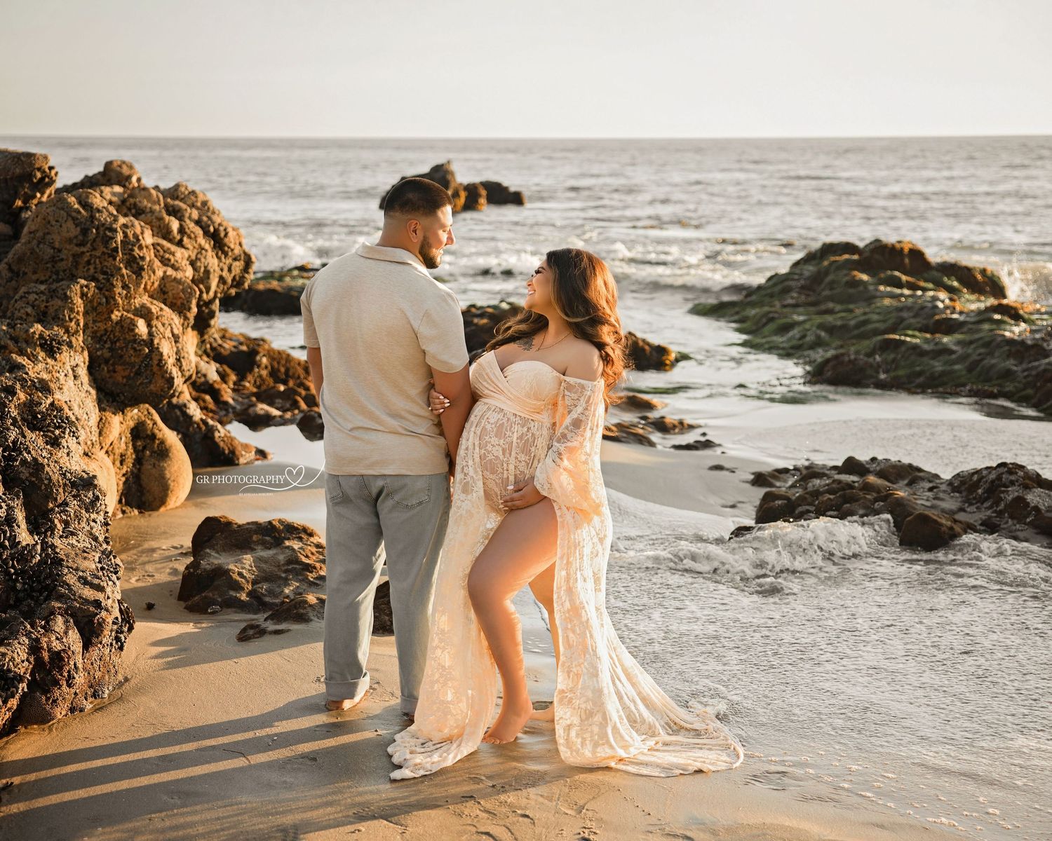 A couple stands together on a rocky beach at sunset, with waves crashing against coastal rocks in the background.