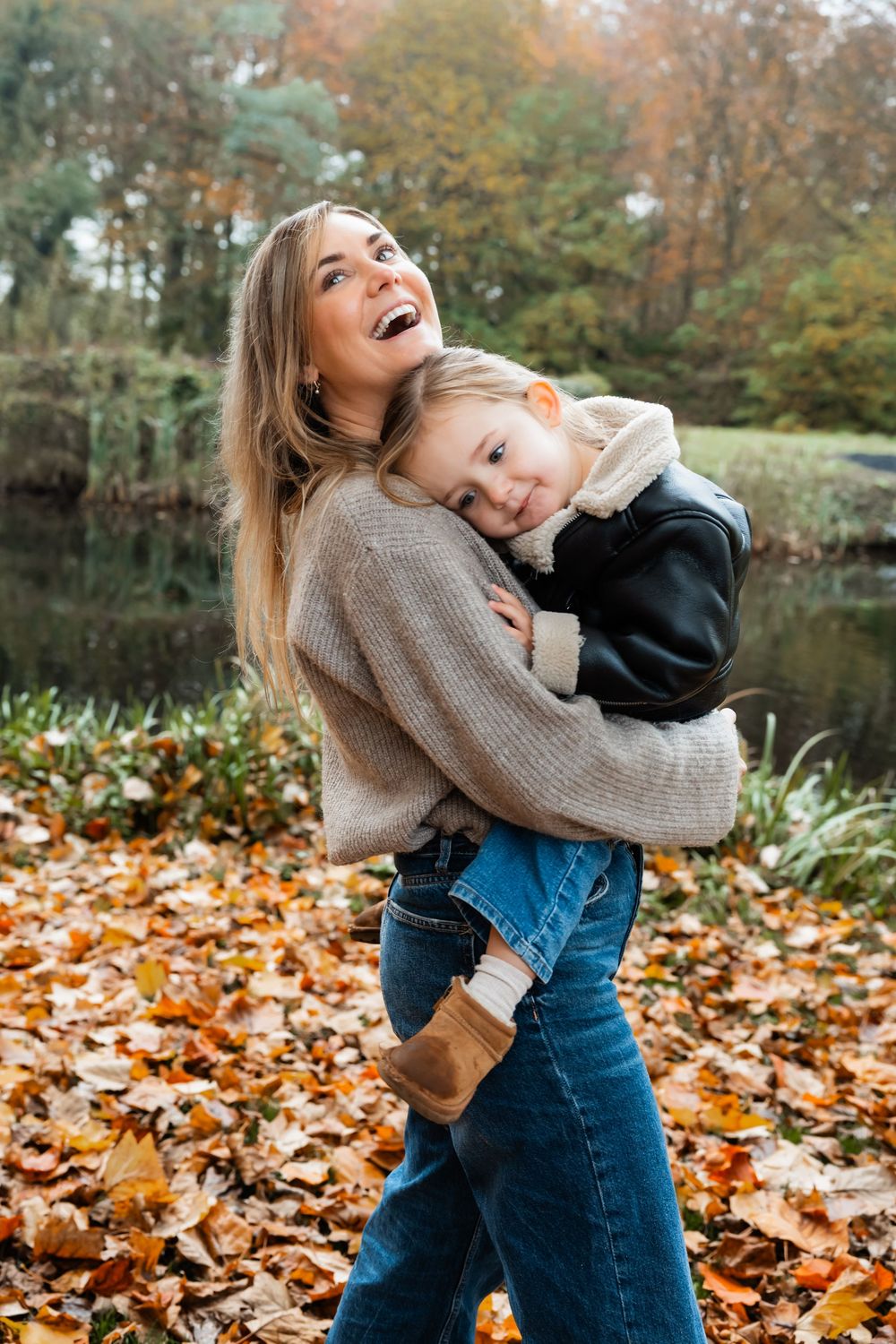 A mother and child play together among fallen autumn leaves by a pond on a crisp fall day.