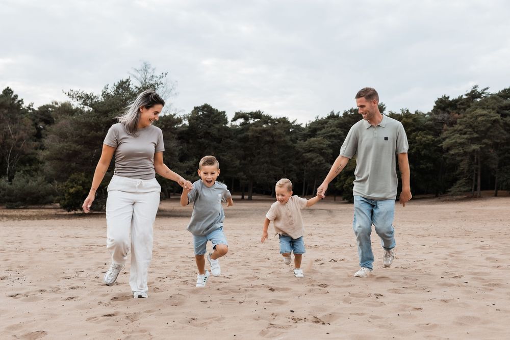 Family in casual clothing walking together and holding hands on a sandy beach with pine trees in background.