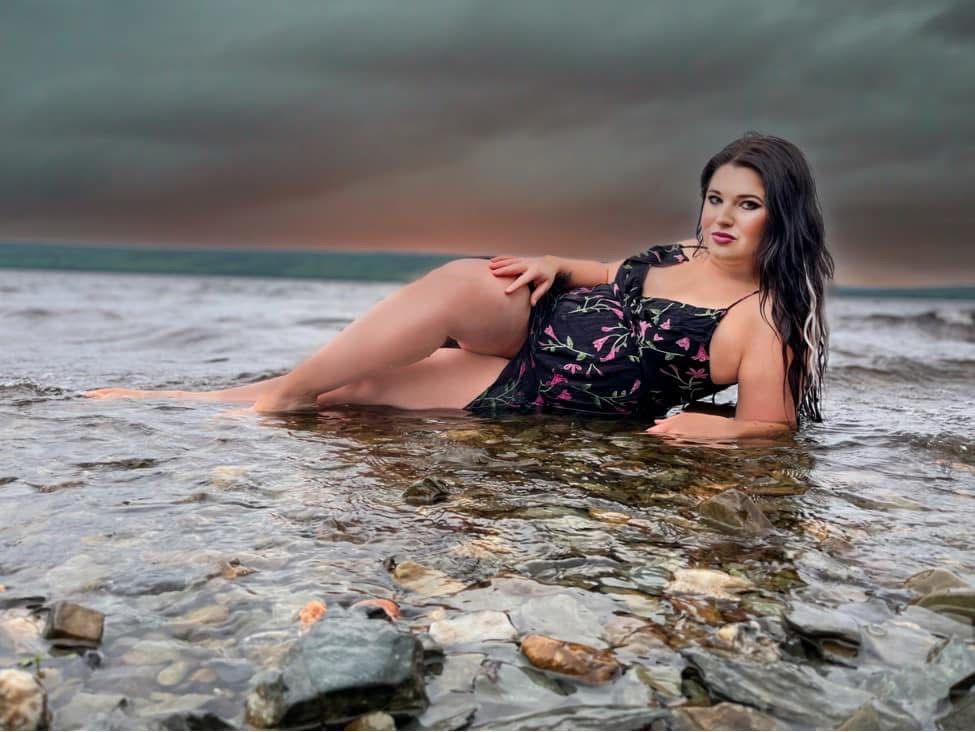 Glamorous woman in floral swimsuit posing dramatically in shallow ocean water under stormy sunset sky.