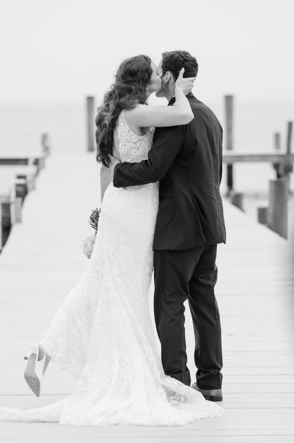 A romantic black and white photo showing a newlywed couple embracing on a dock.