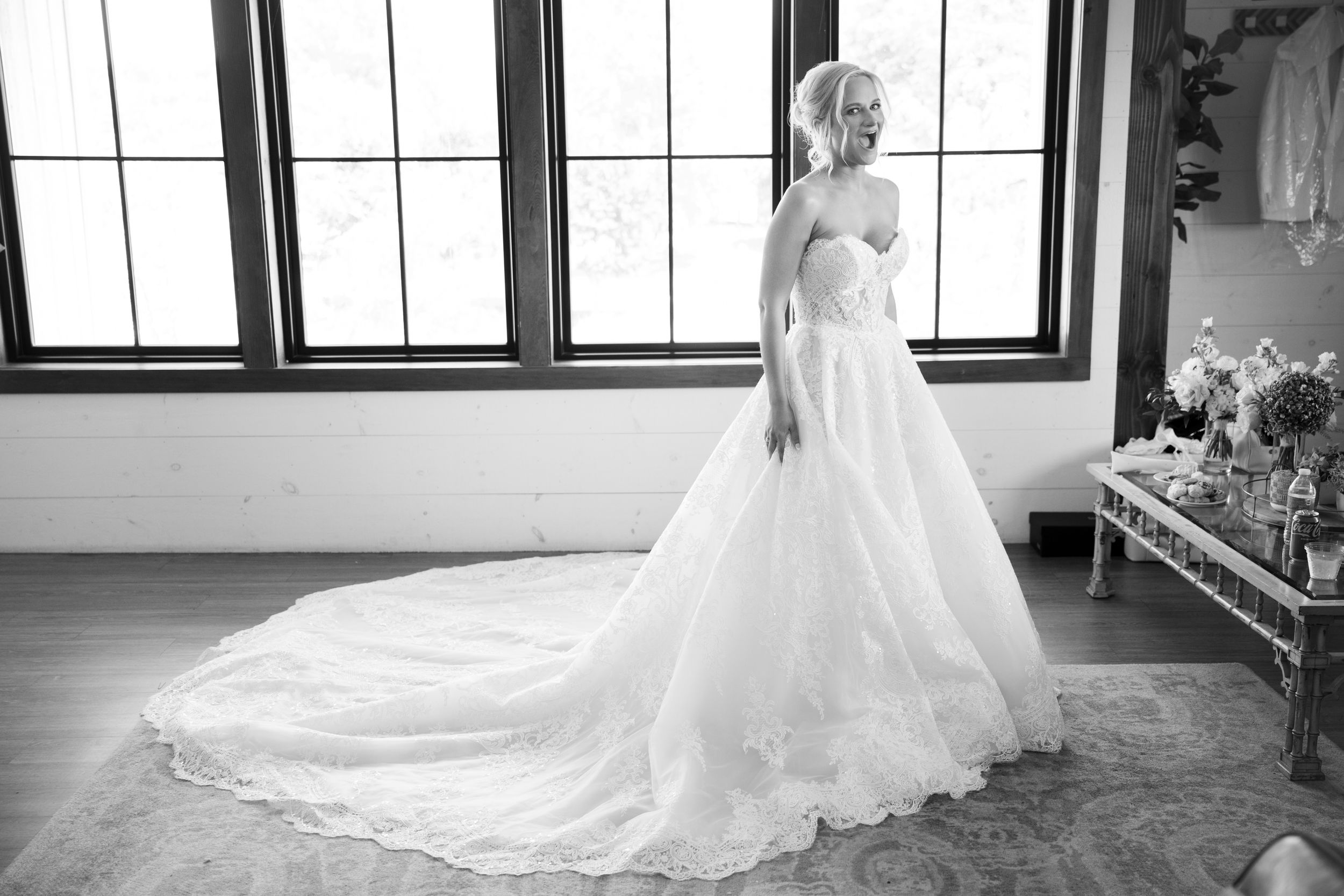 A bride in a flowing white wedding dress with a long train poses in a window-lit room.