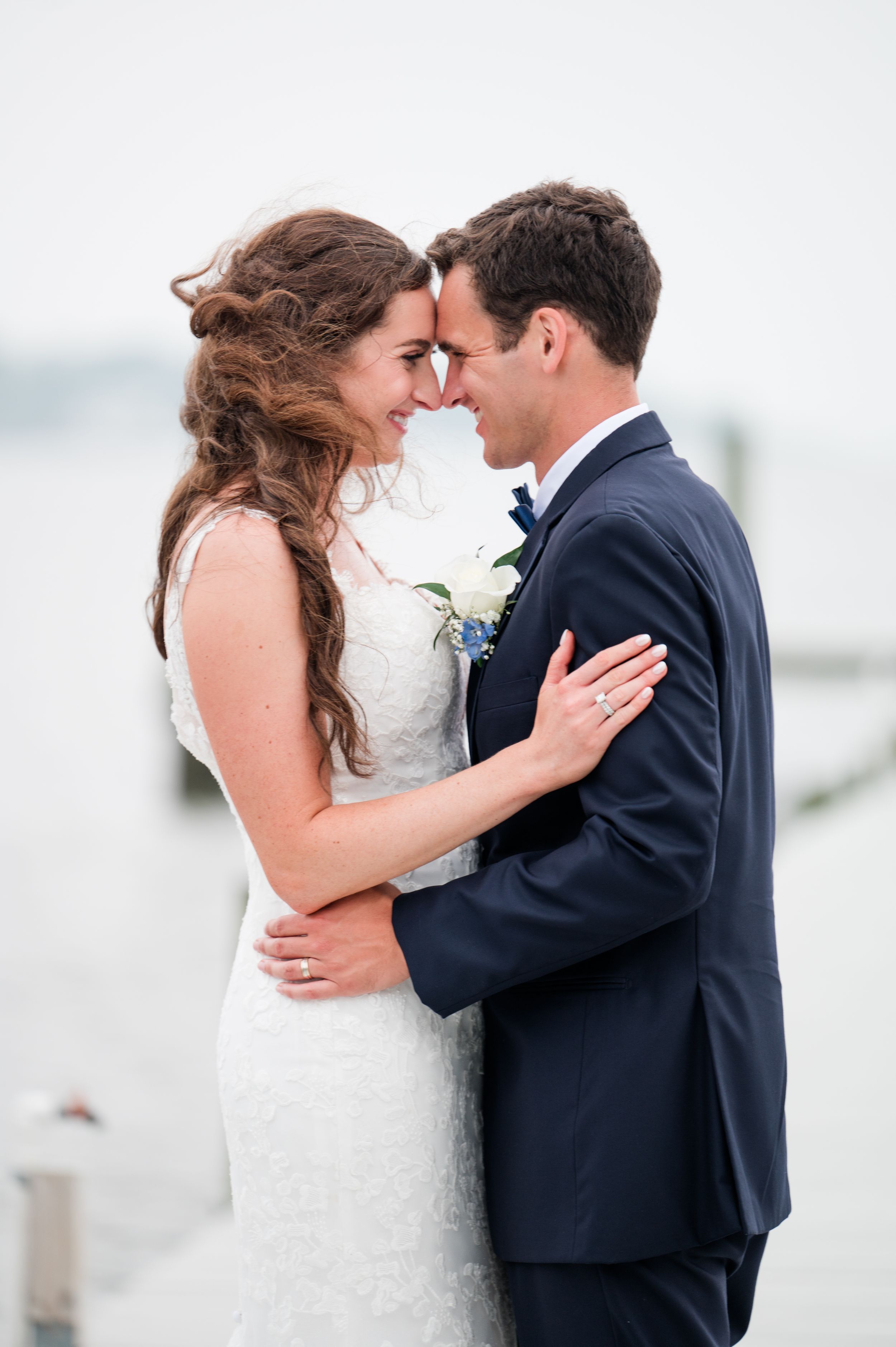 A romantic wedding couple embraces on a cloudy beach day wearing white dress and navy suit.