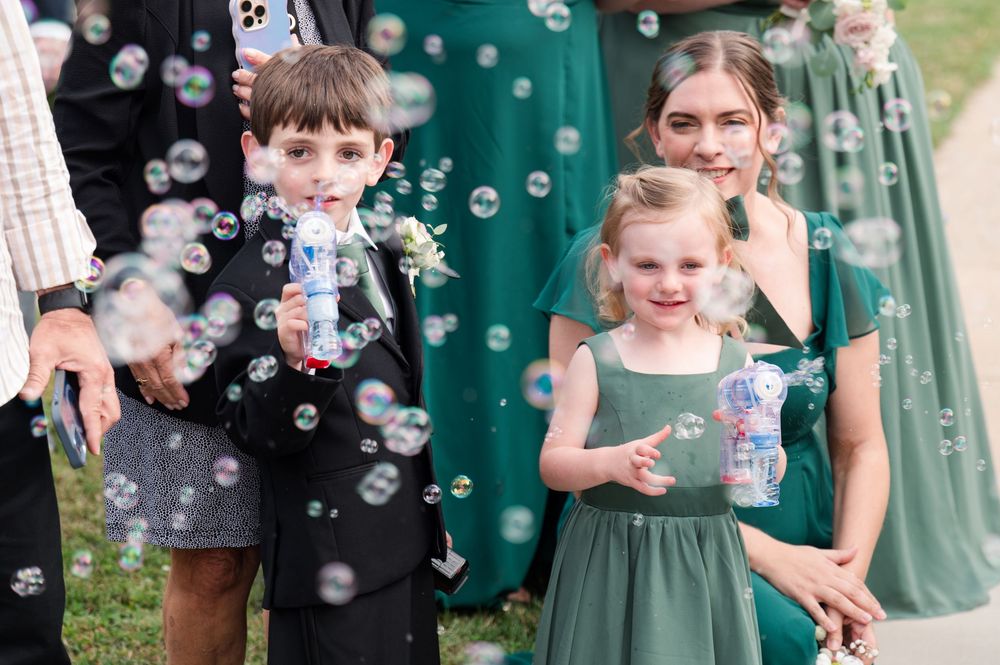 Children in green dresses and a blue suit chase bubbles during an outdoor celebration with a playful atmosphere.