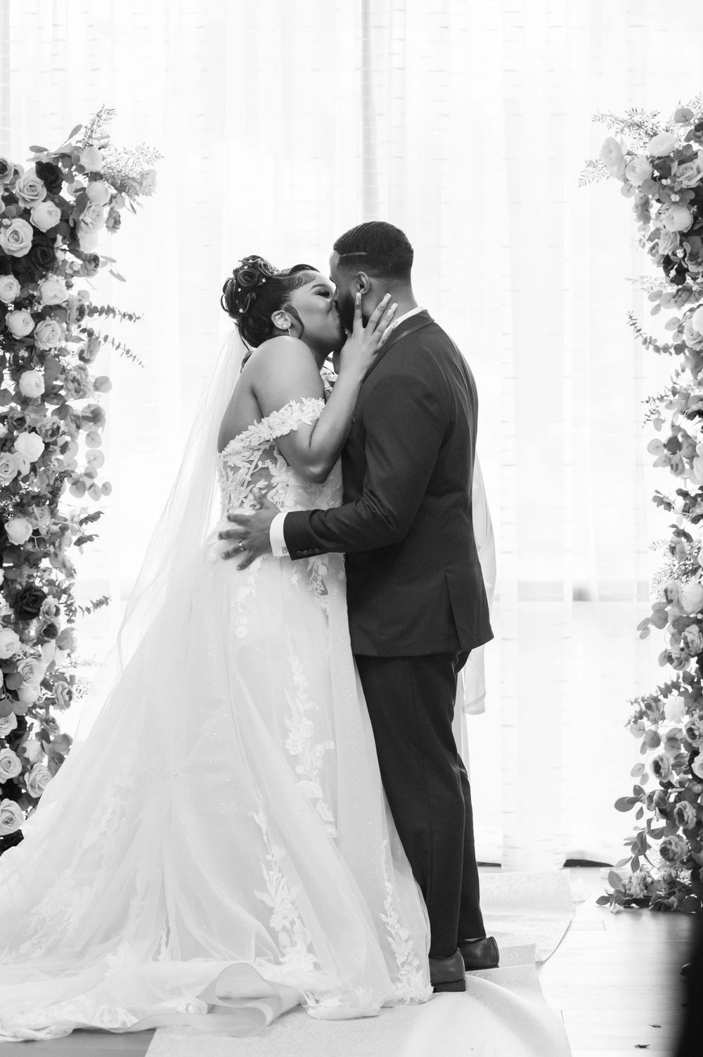 A romantic black and white wedding photo showing a couple kissing under floral arches during their ceremony.
