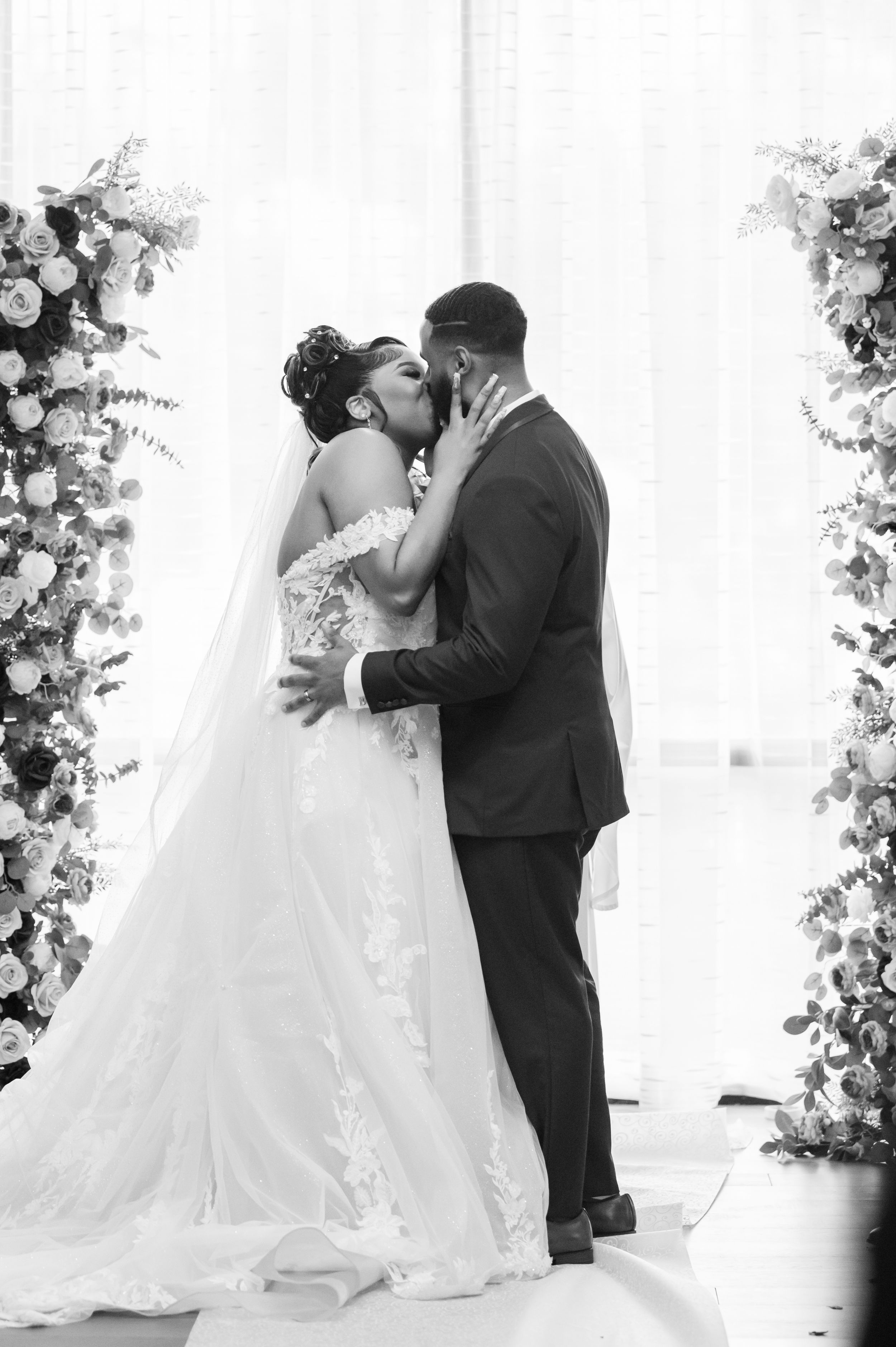 A romantic black and white wedding photo showing a couple kissing under floral arches during their ceremony.