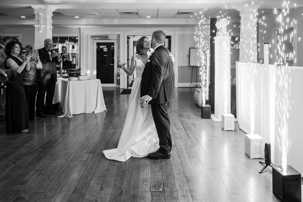 A newlywed couple share their first dance with sparklers in a spacious indoor reception venue with wooden floors while guests look on.