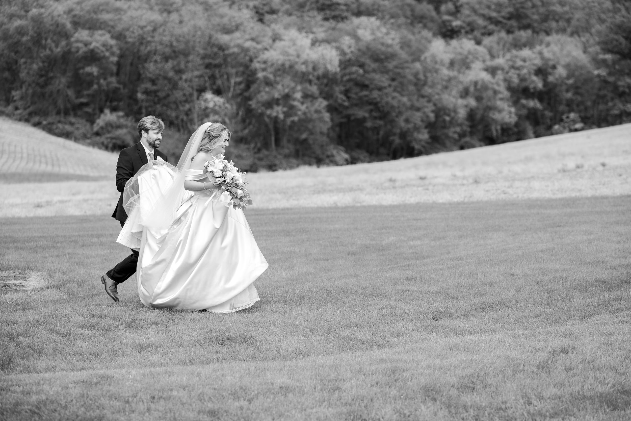 Black and white photo of a bride and groom walking across a grassy field on their wedding day.