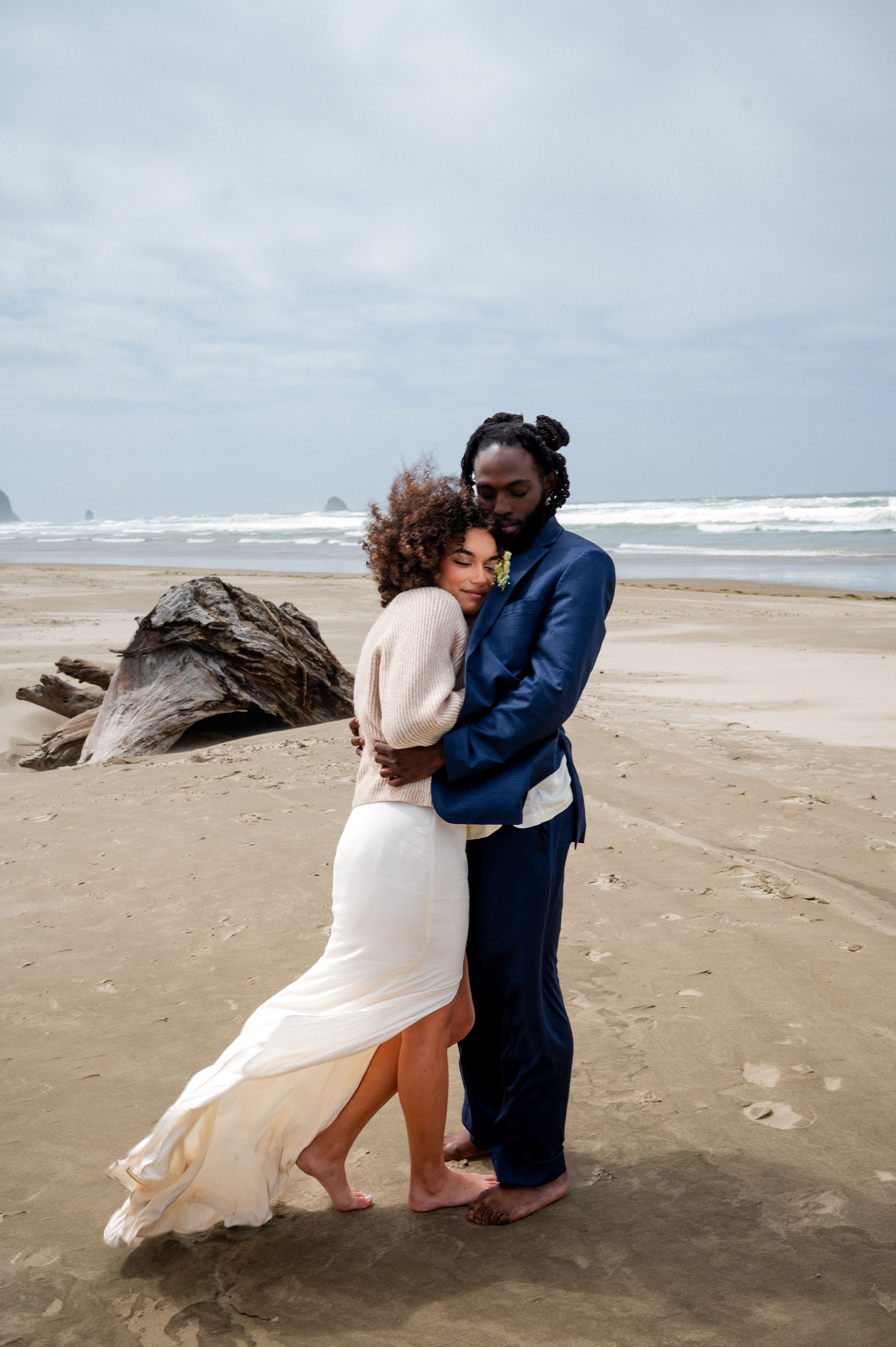 A couple in wedding attire hug on a windswept beach with waves and driftwood in the background.