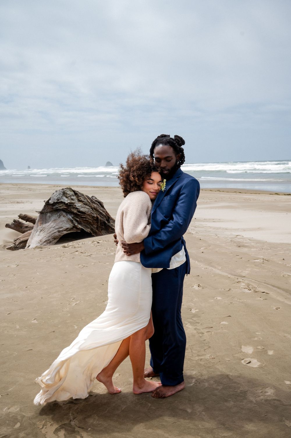 A couple in wedding attire hug on a windswept beach with waves and driftwood in the background.