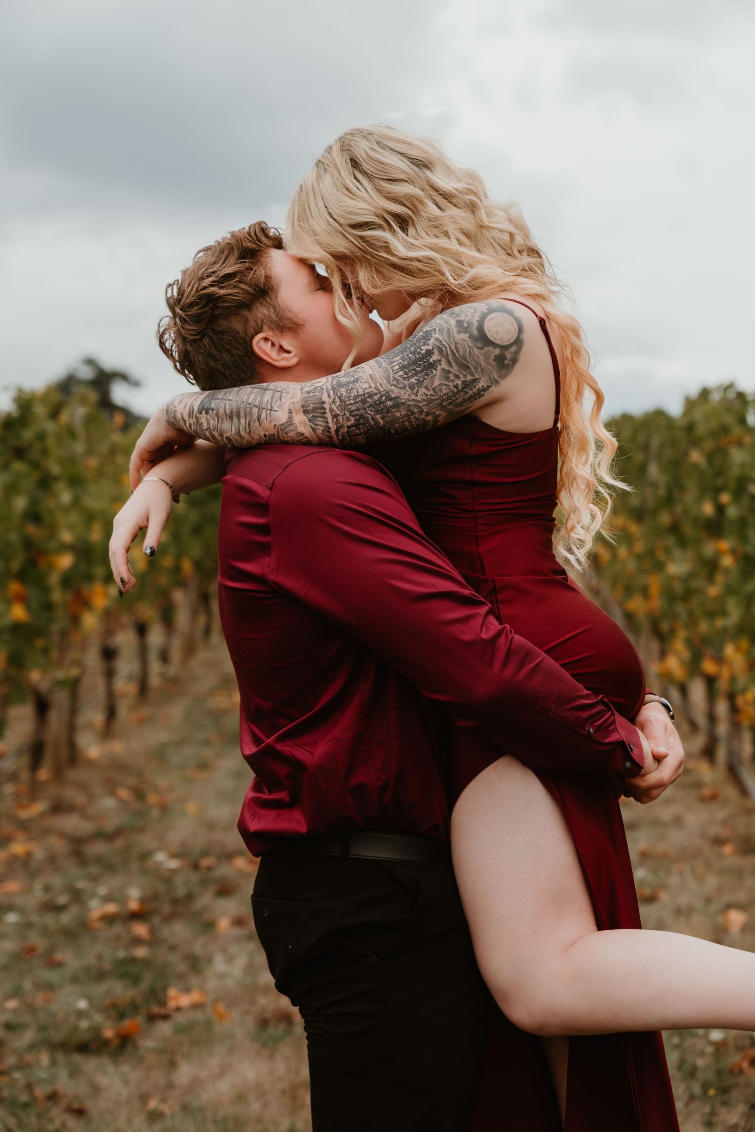 Couple kissing passionately in a vineyard, woman in red dress lifted by tattooed man in burgundy shirt.