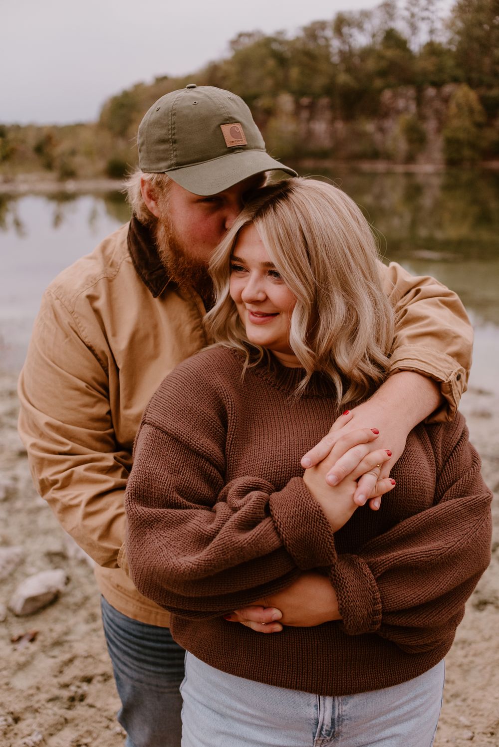 Affectionate couple laughing and hugging by a lake in warm earth-toned sweaters.