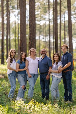 Group of people wearing coordinated denim and white outfits pose together in a sunny forest setting with wildflowers.