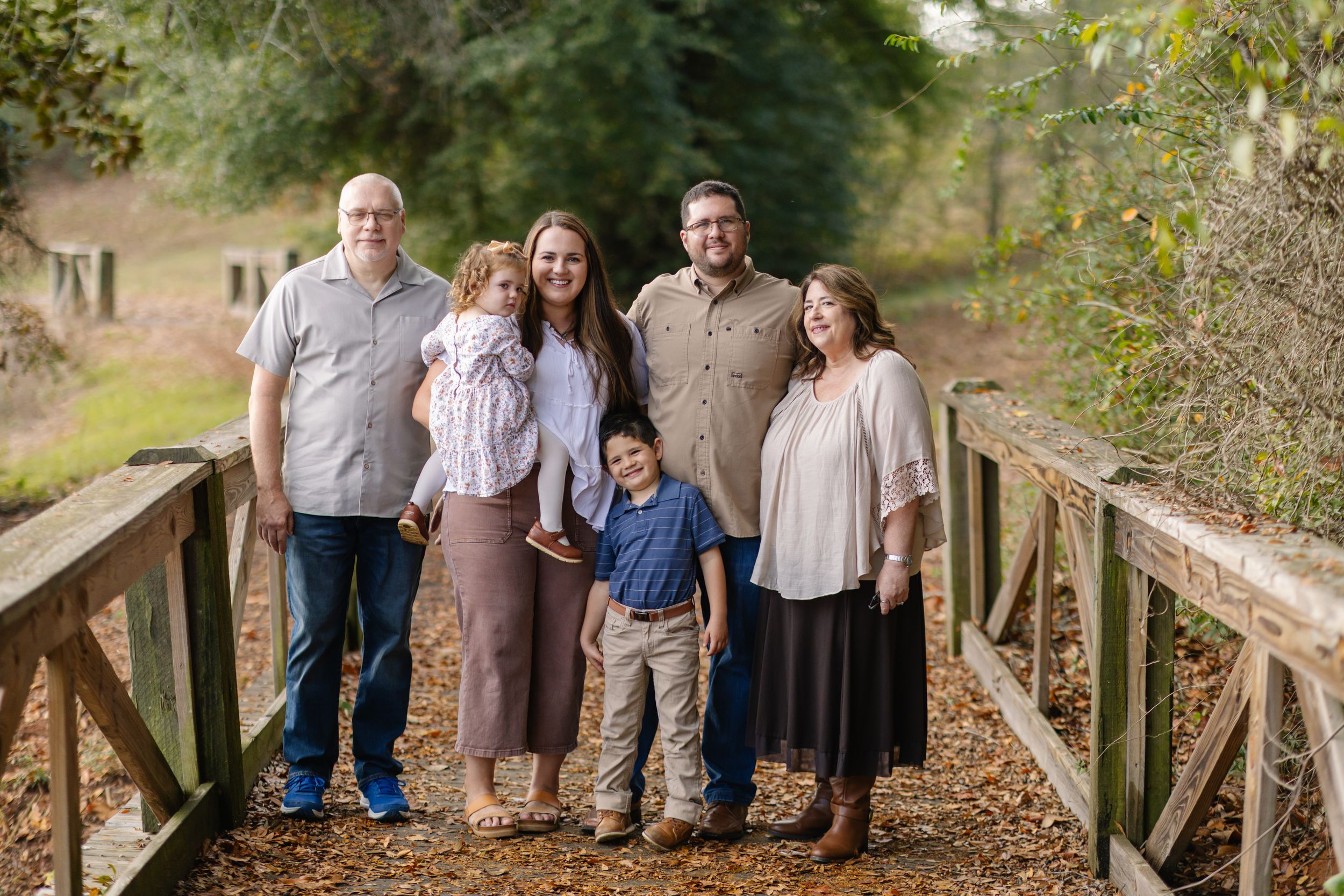 Family group stands together on wooden bridge surrounded by autumn foliage in outdoor setting.