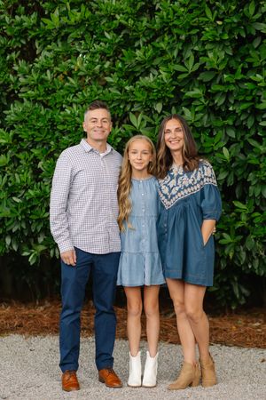 Three siblings pose together in coordinating blue and white outfits against a lush green hedge backdrop.