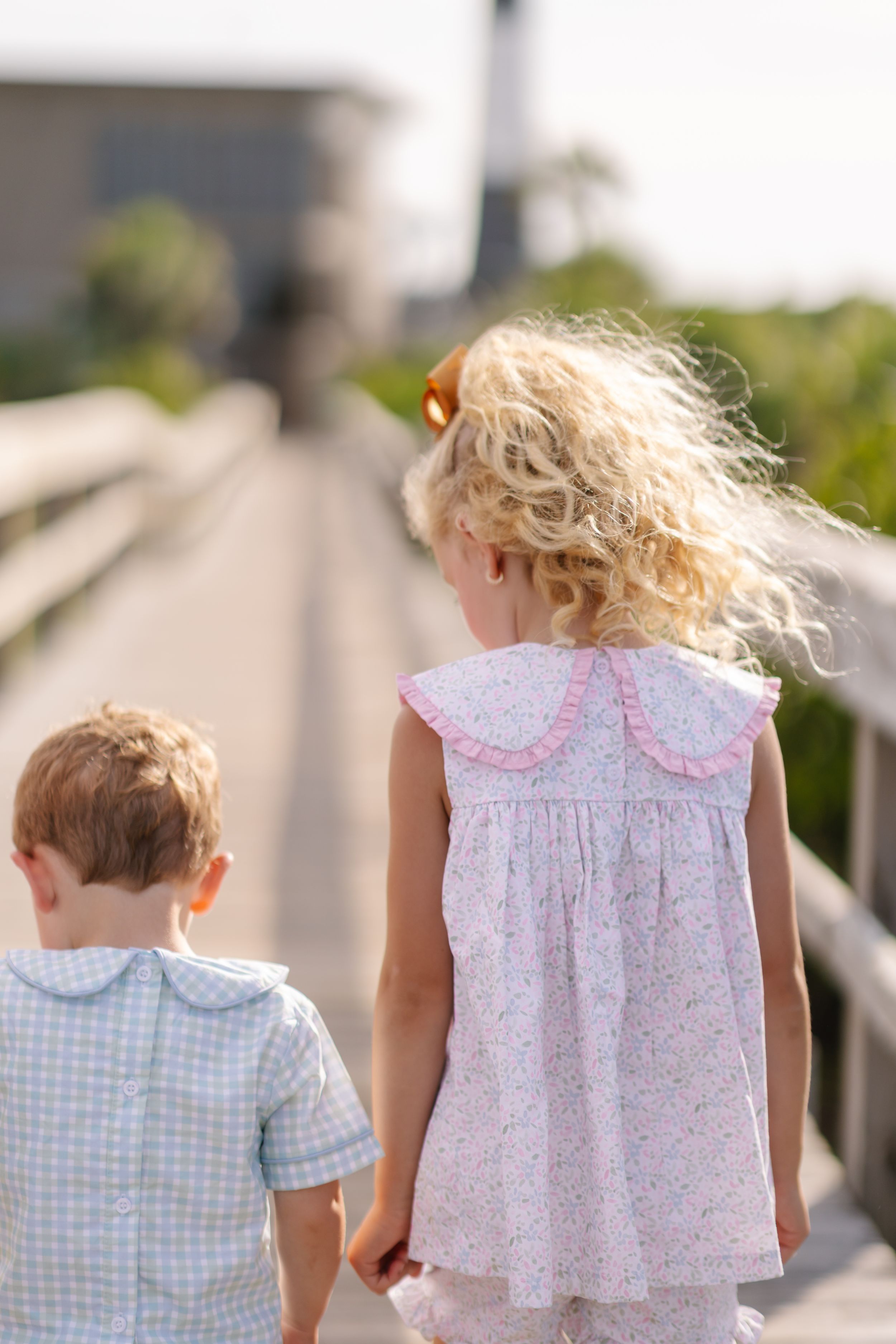 Two young children in light summer clothing walk together along a railing on a sunny day.