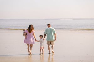 A family walks together holding hands along a serene beach at sunset, leaving footprints in the wet sand.