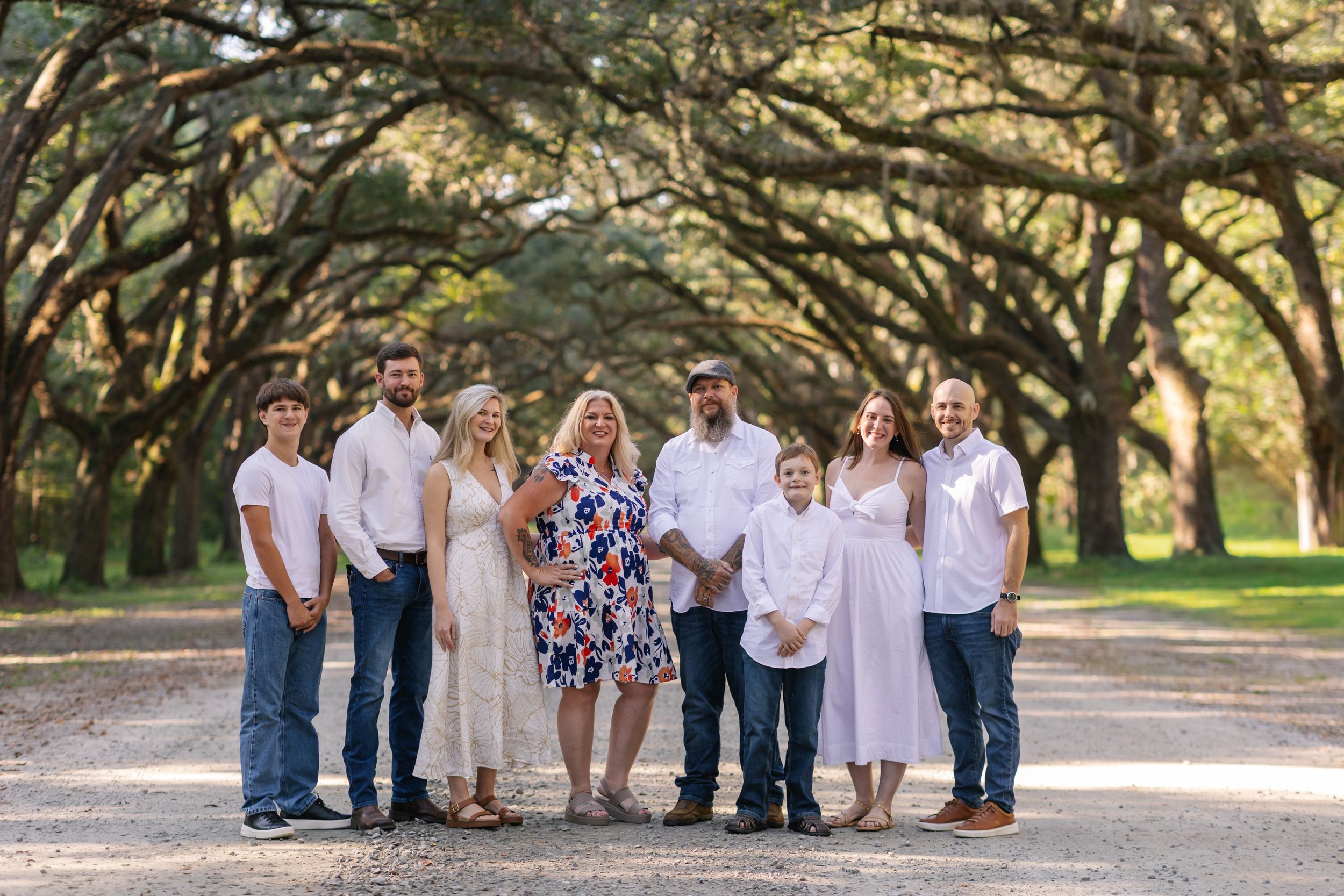 A group of people in coordinating white and blue outfits pose together for outdoor family photos under oak trees.
