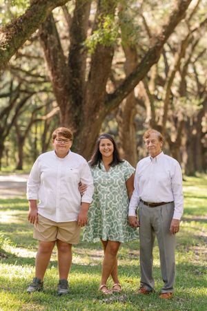 A family of three poses together in a grassy park setting with oak trees in the background.