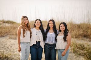 Four friends in casual denim outfits pose together on a sandy beach path at sunset.