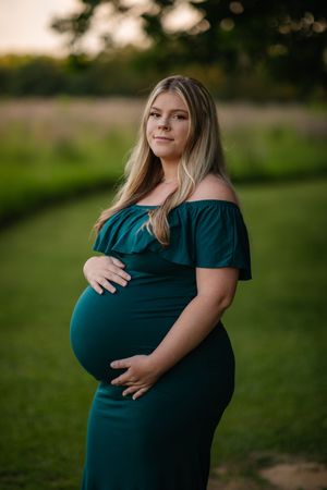 Pregnant woman in dark teal dress poses outdoors during golden hour maternity photoshoot.