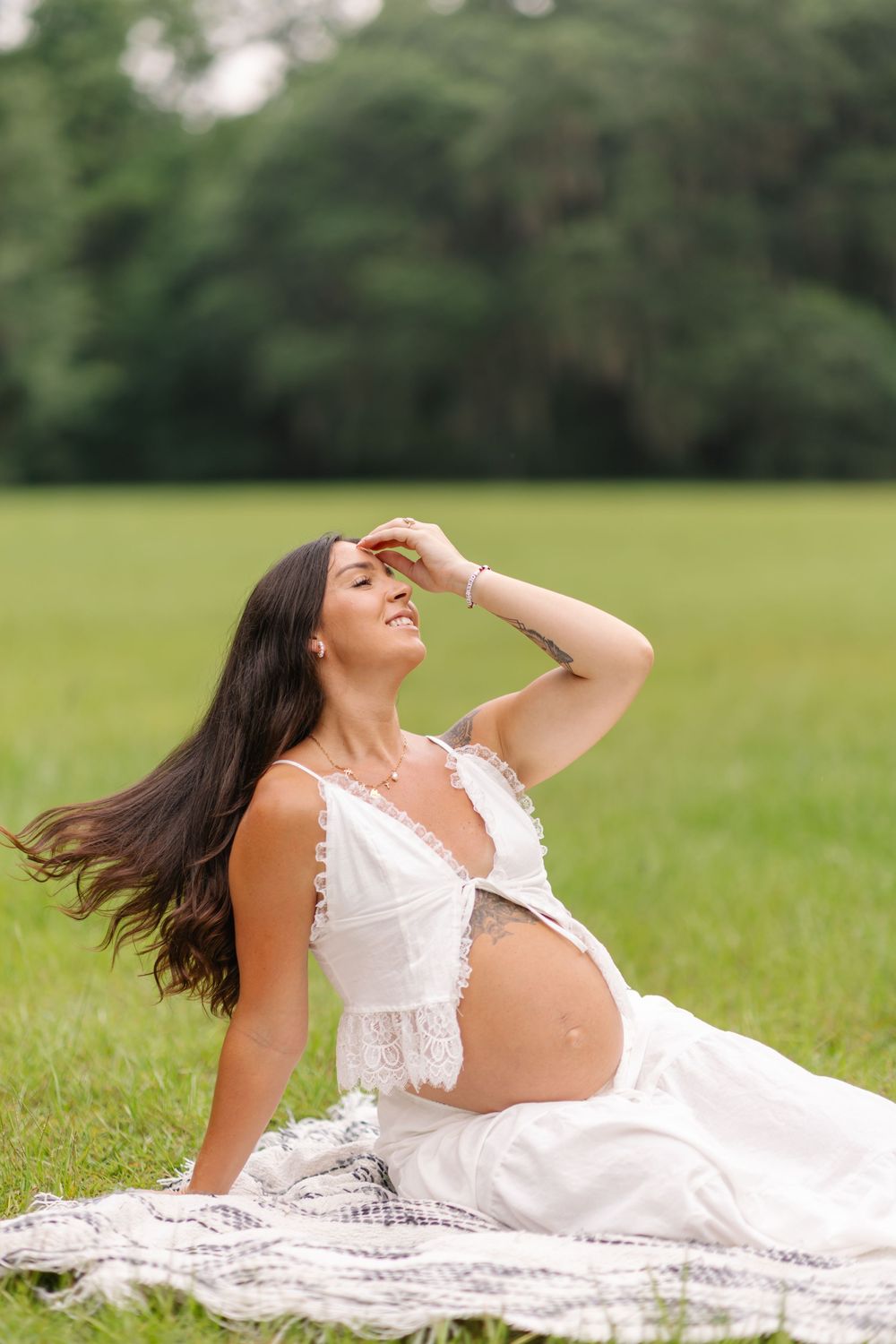 Pregnant woman in white lace crop top sits on blanket in green field, hair blowing, hand shielding eyes from sun.