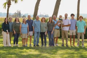 A group of people standing together on grass with palm trees and ocean in the background on a sunny day.