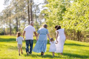 Family of five walking hand-in-hand through a sunny green park surrounded by tall pine trees, dressed in white outfits.