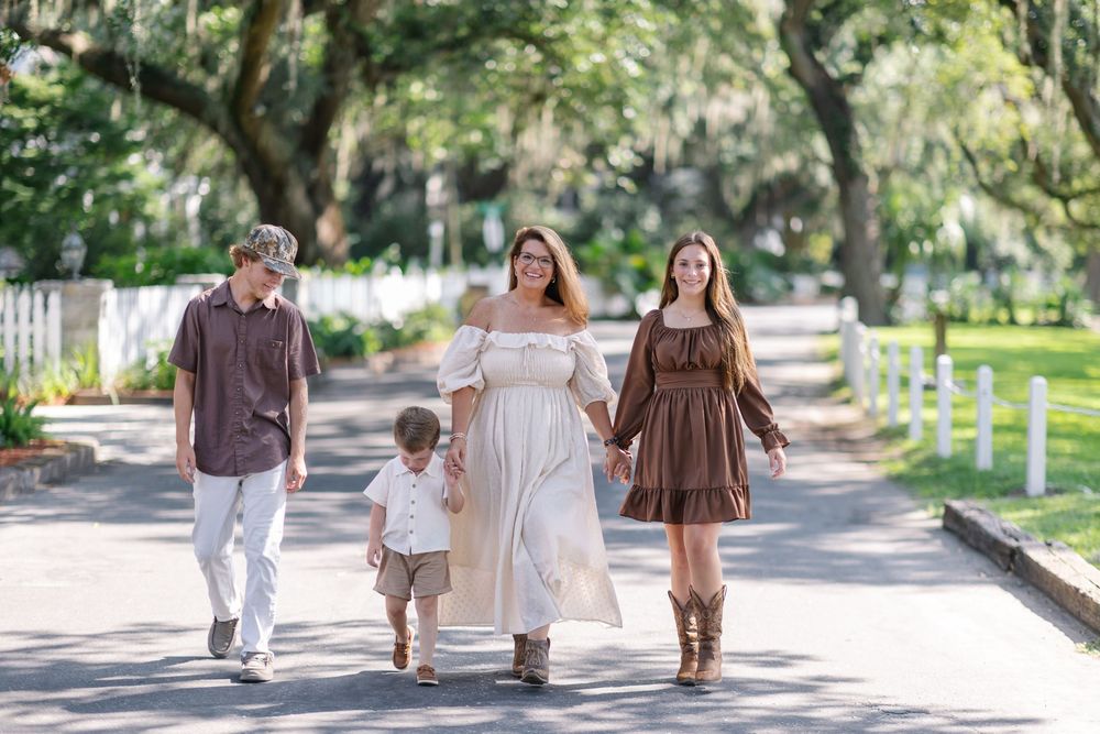 Family of four walking down a tree-lined path, mom in white dress, daughter in brown dress, son in maroon shirt.