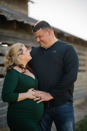 Pregnant couple sharing a tender moment outdoors, woman in green dress, man in black Under Armour shirt, rustic wood backdrop.