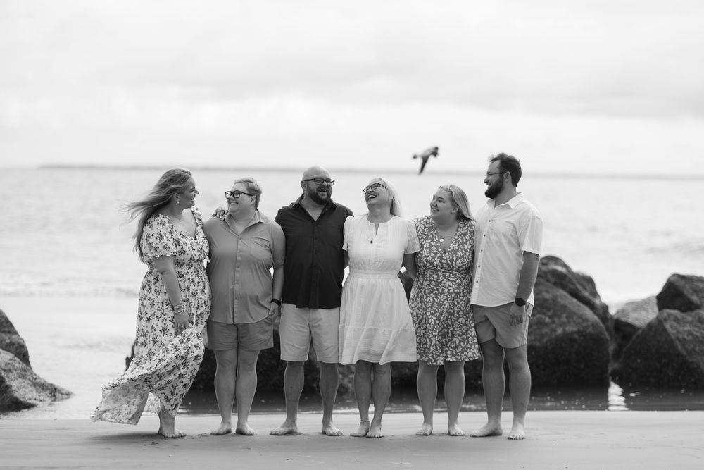 Black and white photo of six adults laughing together on a rocky beach with ocean waves in the background.
