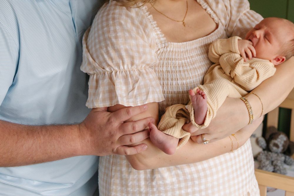 Parents cradling a sleeping newborn baby, mother in a white dress gently holding infant with father's hand nearby.