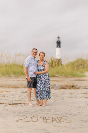 A couple stands together on a sandy beach with '20 years' written in the sand and a lighthouse in the background.