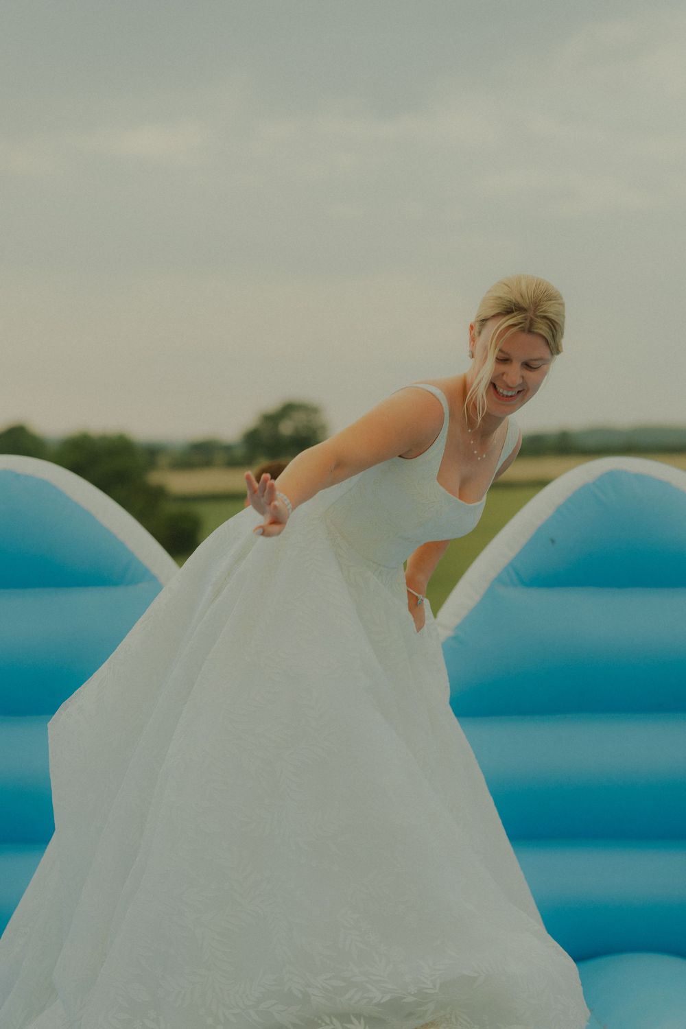 Bride in white wedding gown laughing on a blue bouncy castle outdoors.