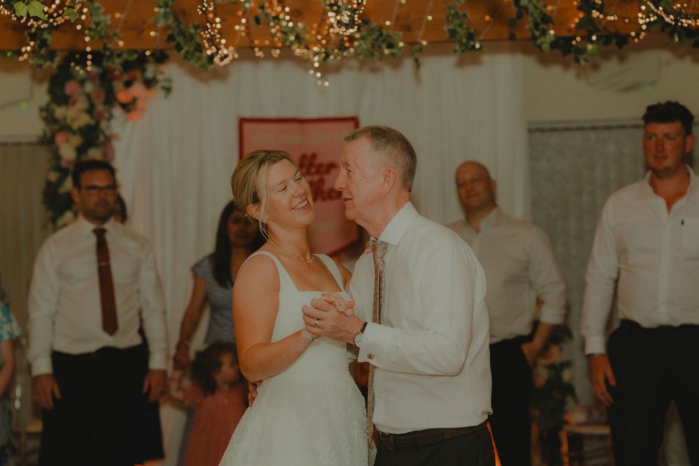Bride and groom share their first dance, smiling joyfully, surrounded by guests at a decorated wedding reception.