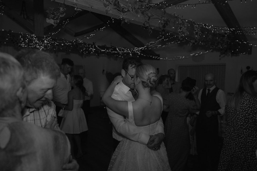 Black and white photo of bride and groom sharing first dance at wedding reception with fairy lights overhead.