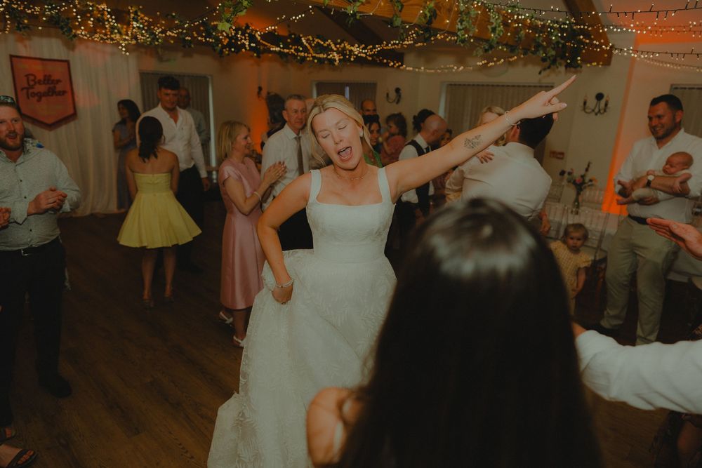 Bride in white gown dancing joyfully with guests at lively wedding reception with warm lighting.