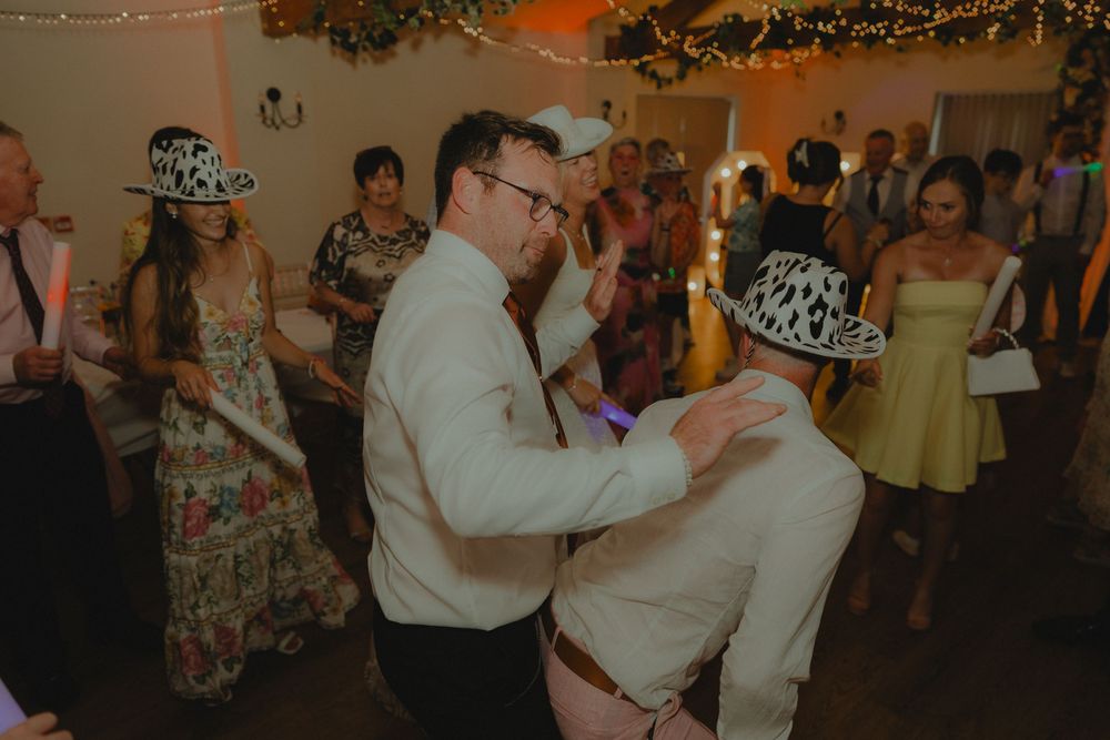 Guests dancing joyfully at a wedding reception, wearing cowprint hats in a warmly lit venue.