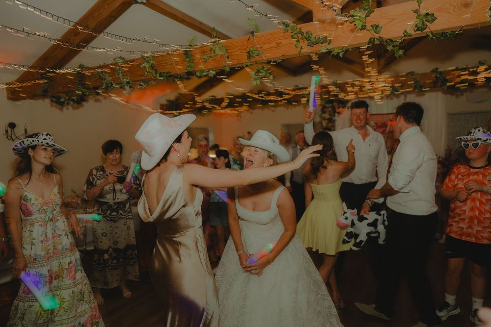 Two brides in white hats dancing joyfully with guests at a rustic wedding reception venue.