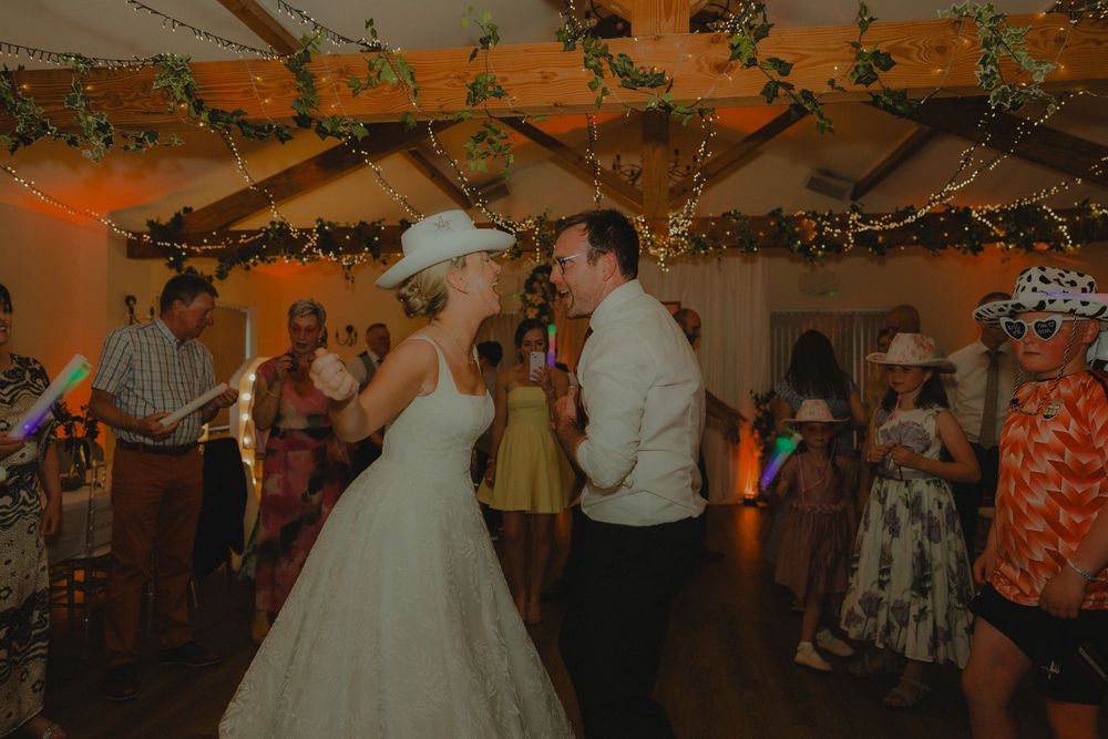 Bride and groom share first dance in cowboy hats at rustic wedding reception with fairy lights.