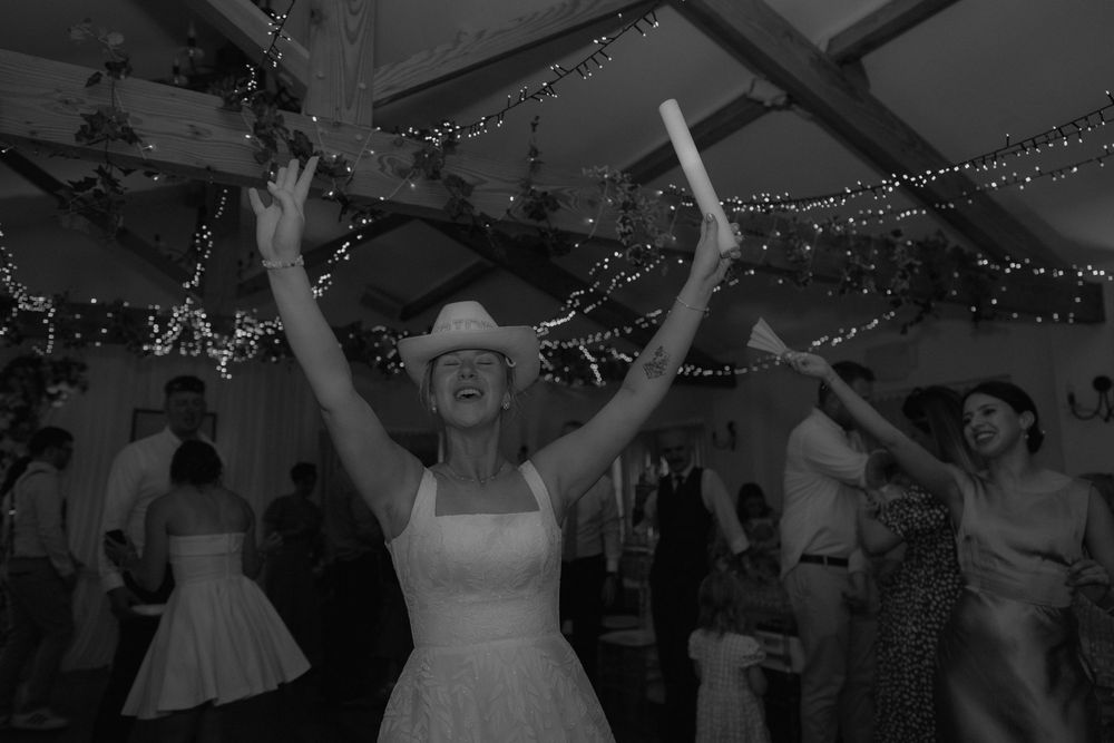 Bride in cowboy hat dancing joyfully with arms raised at wedding reception with fairy lights.