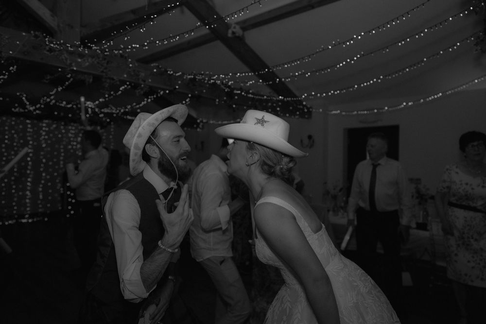 Black and white photo of couple dancing in cowboy hats at wedding reception with fairy lights.