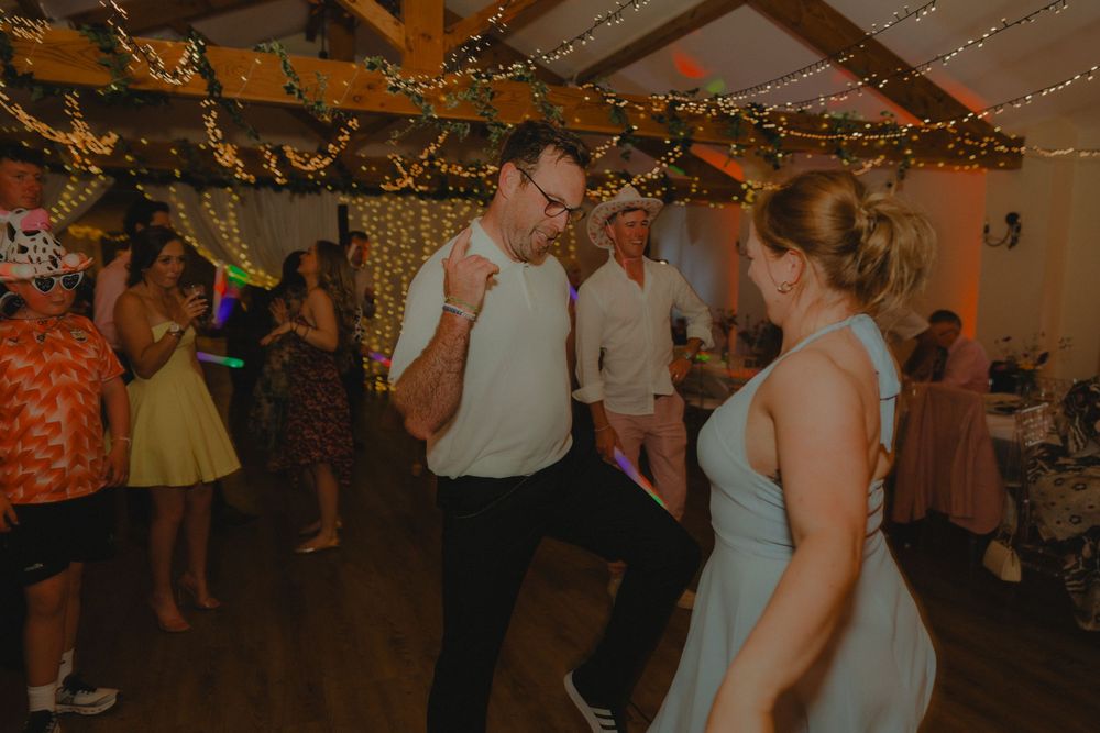 Guests dancing joyfully at rustic wedding reception venue decorated with fairy lights and greenery.