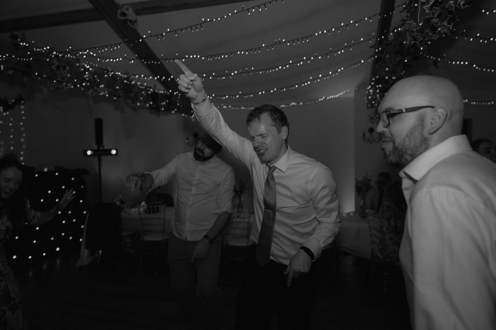 Black and white photo of men dancing and celebrating at a wedding reception under fairy lights.