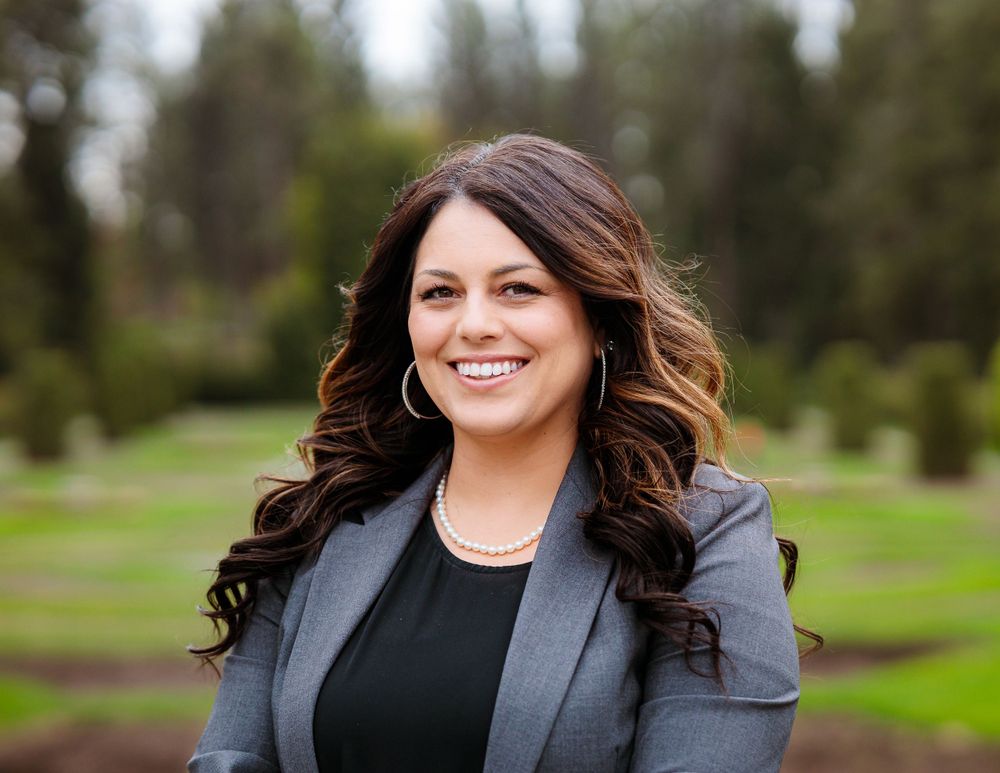 A professional woman in a suit and black shirt stands confidently in front of a lush Manito park backdrop.